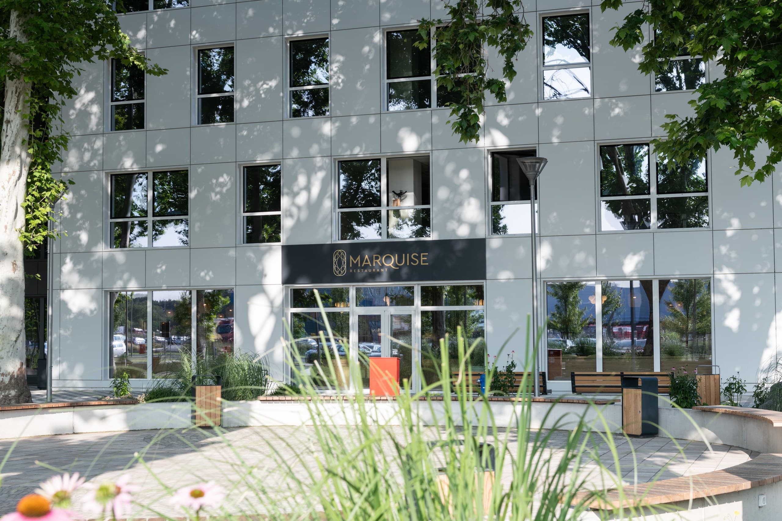 Modern white building facade featuring Marquise Restaurant with large glass windows and lush green landscaping.