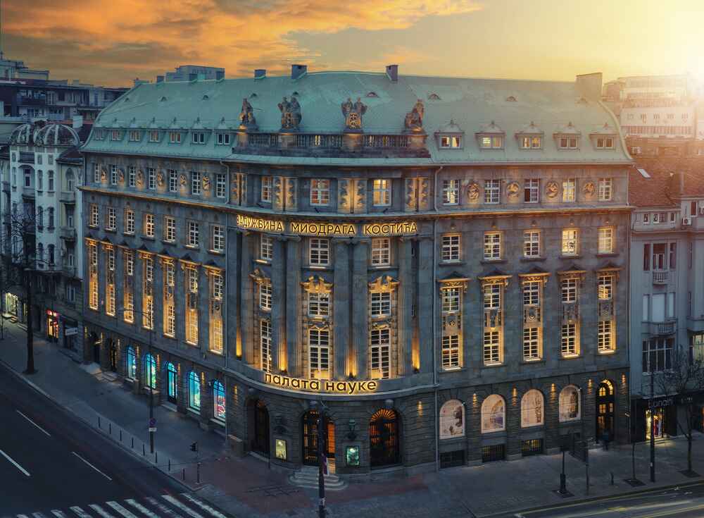 Ornate stone building with illuminated windows and Cyrillic text at dusk under a sunset sky.